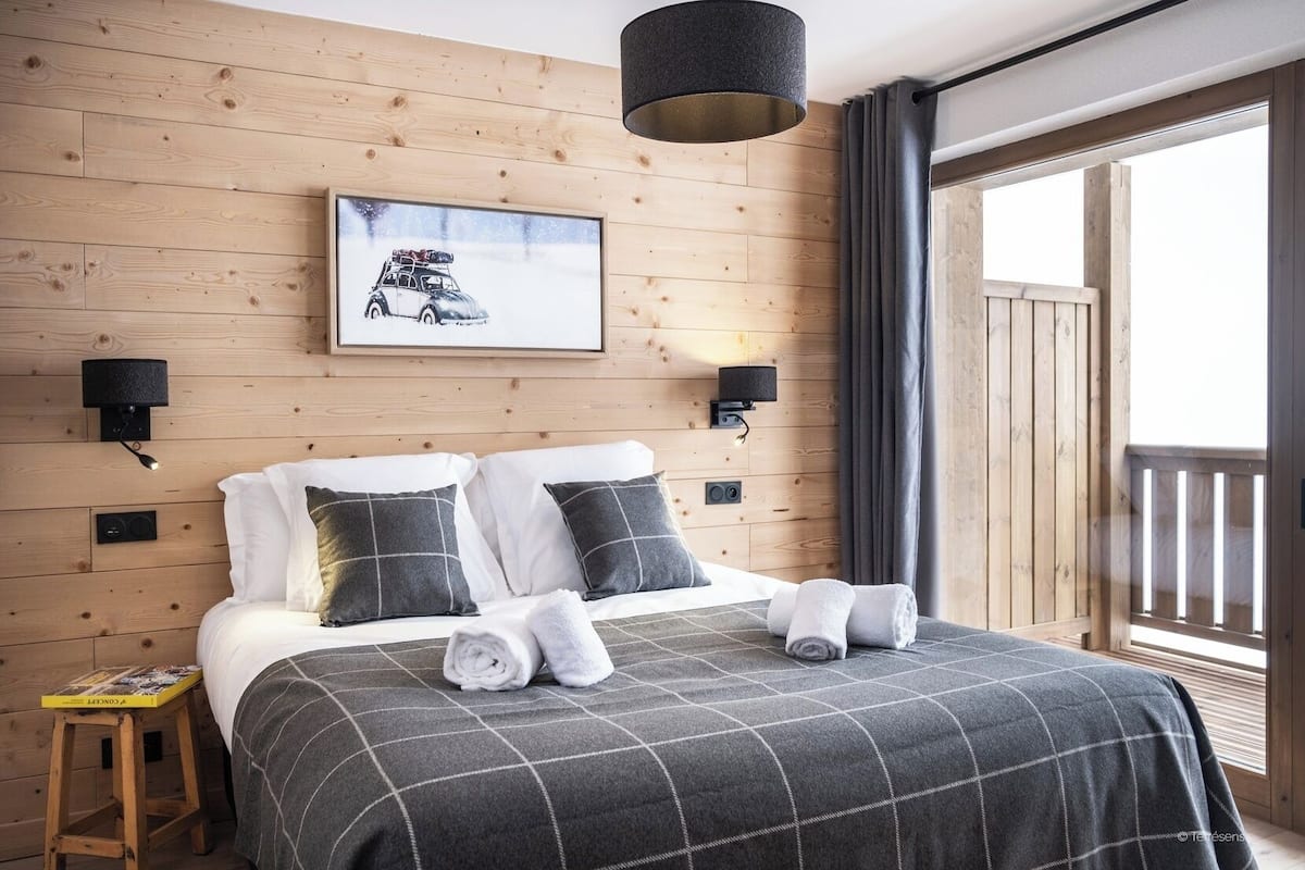 Ein Hotelzimmer mit Holzwand, einem Bett mit kariertem Überwurf, zwei Kissen, einem gerahmten Bild über dem Bett, einem Nachttisch mit einem Buch und einem Balkon mit Vorhängen.