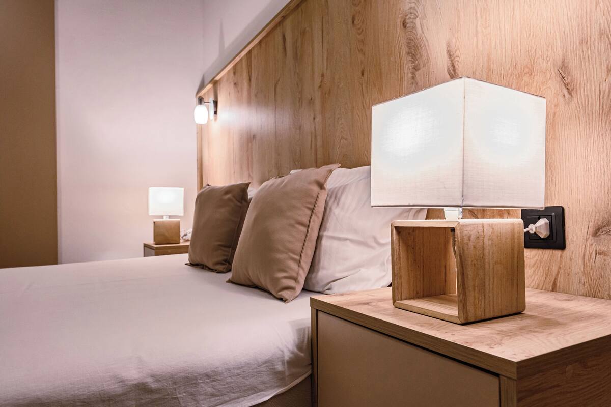 Ein ordentlich bezogenes Bett mit beige farbenem Bettzeug, ein Holzkopfbrett, ein Nachttisch mit Lampe und ein Holznachtschränkchen.