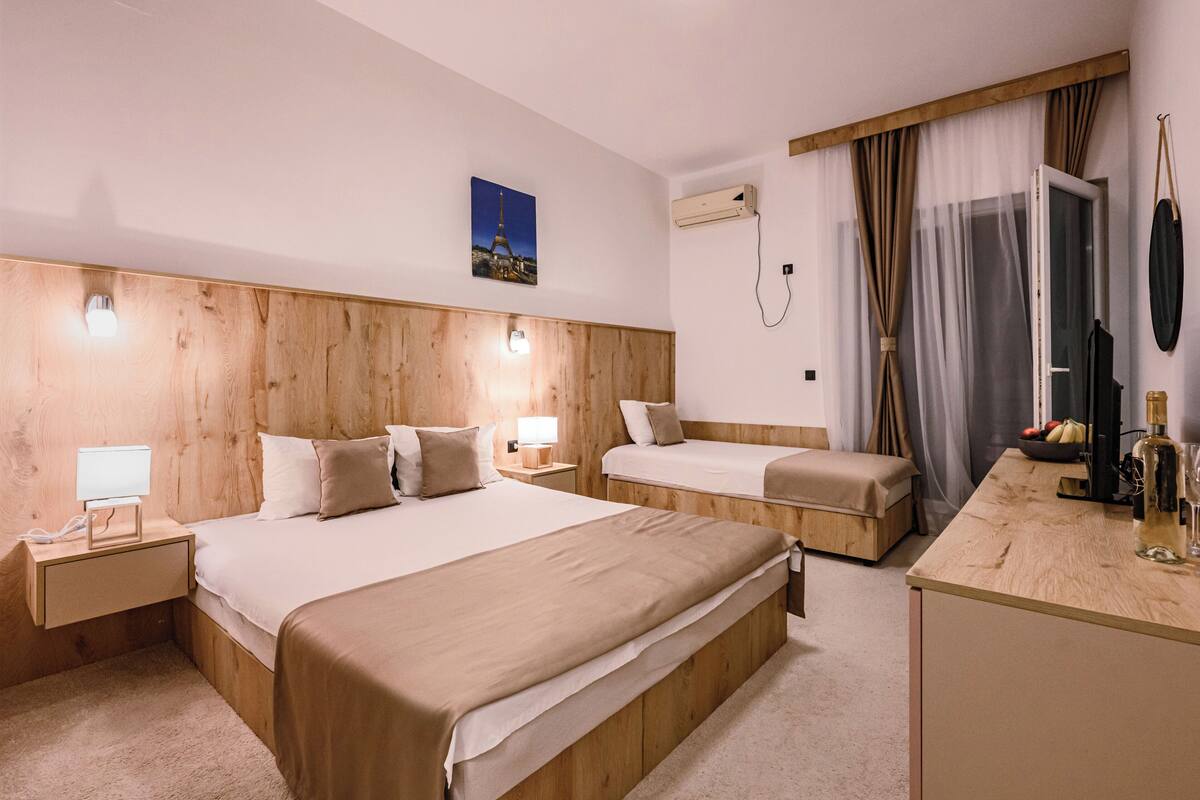 Ein Hotelzimmer mit einem hölzernen Kopfteil, zwei Betten, einem Nachttisch mit Lampe, einem kleinen Schreibtisch mit Flasche und Gläsern sowie einem Bild des Eiffelturms an der Wand.