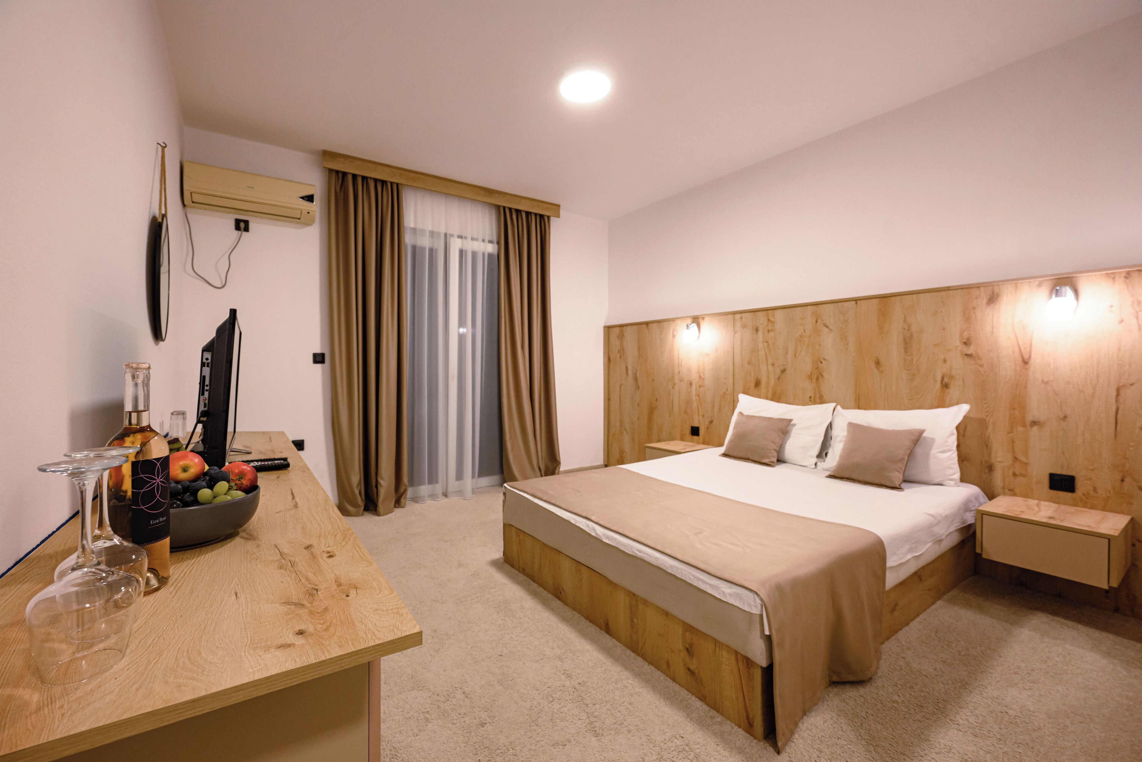 A hotel room with a wooden bed, a bedside table, a desk with a bowl of fruit and a bottle, and an air conditioning unit.