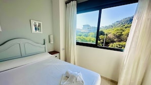 A neatly made bed with white linens and towels, a bedside table with a lamp, and a window with curtains offering a view of greenery and buildings.