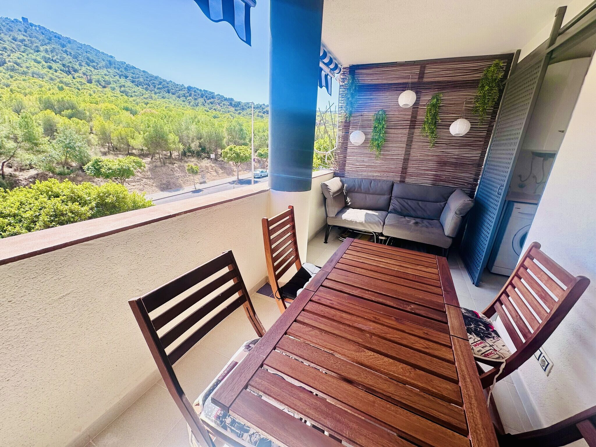 A balcony with a wooden table and chairs, a sofa, and a view of a green landscape.