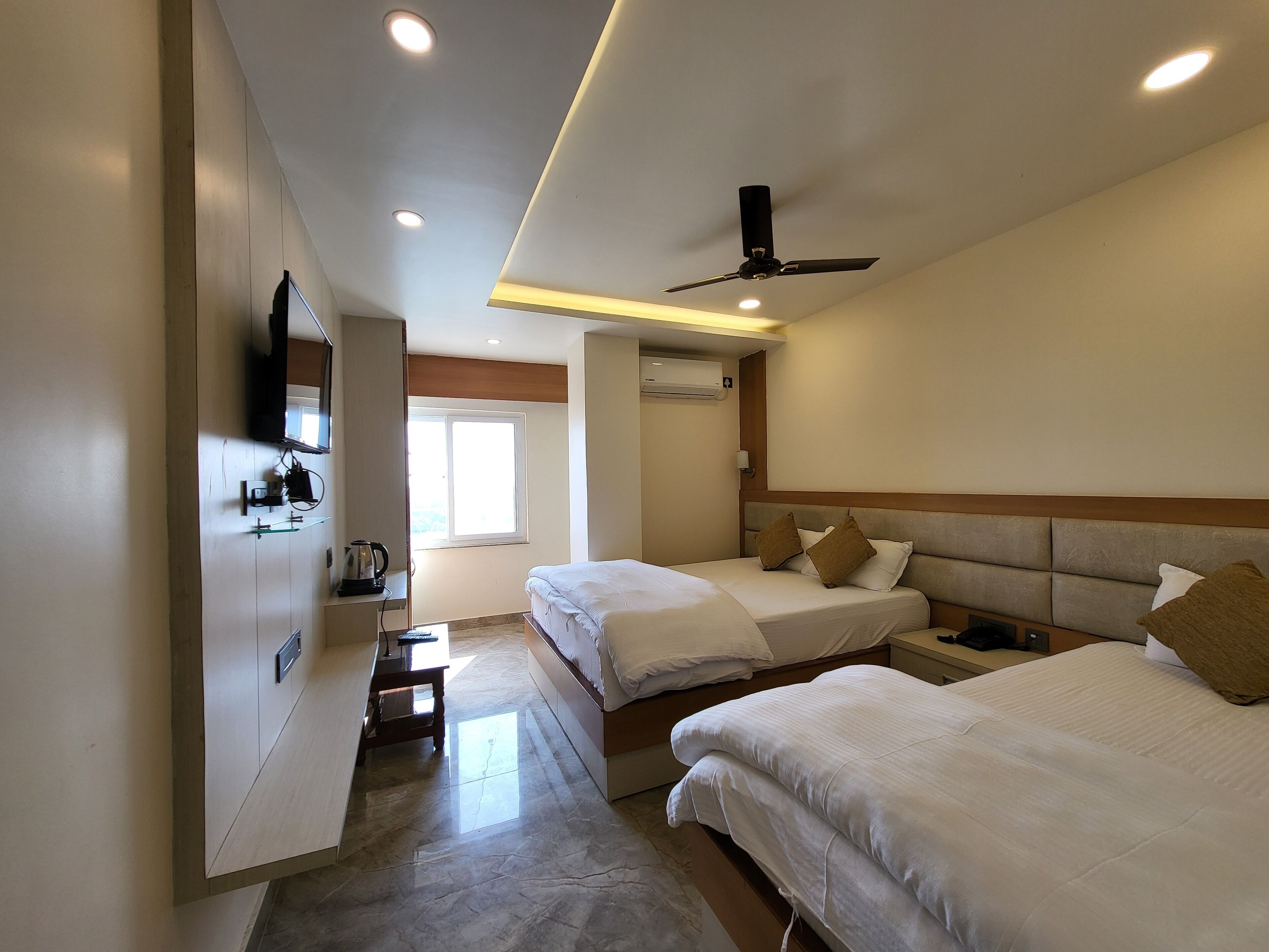 Una habitació d'hotel amb dos llits, un ventilador de sostre, un televisor fixat a la paret i una finestra que deixa entrar llum natural.
