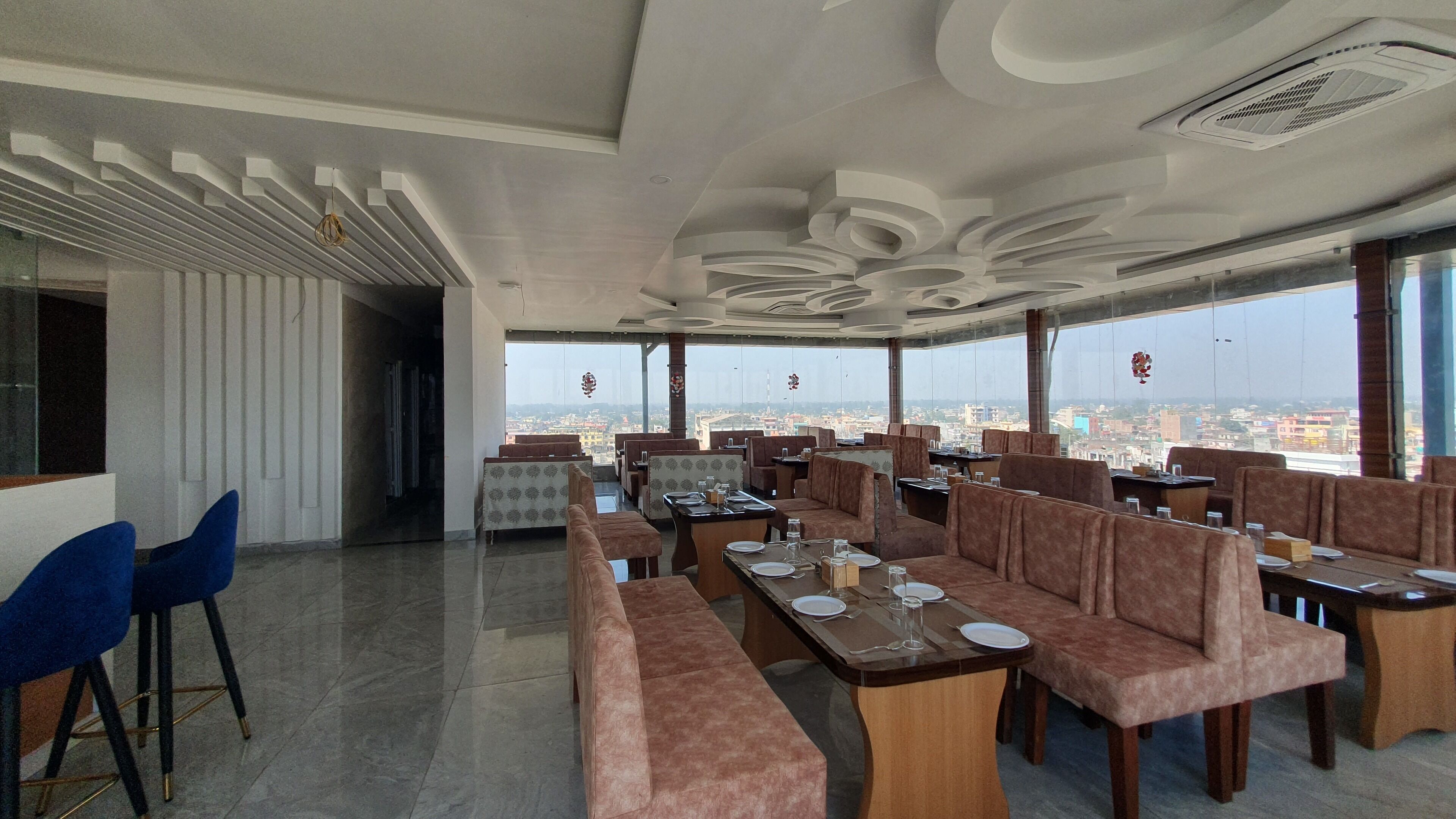 Un restaurante con mesas altas y cabinas, grandes ventanales con vistas a la ciudad y un diseño moderno del techo.