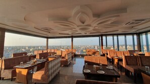Restaurante com teto alto e vista para a cidade. Mesas montadas com copos e talheres.