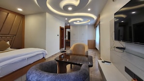 A modern hotel room with a bed, a seating area with a table and chairs, a TV mounted on the wall, and a wardrobe with sliding doors. - Anjani Suite (Janakpur)