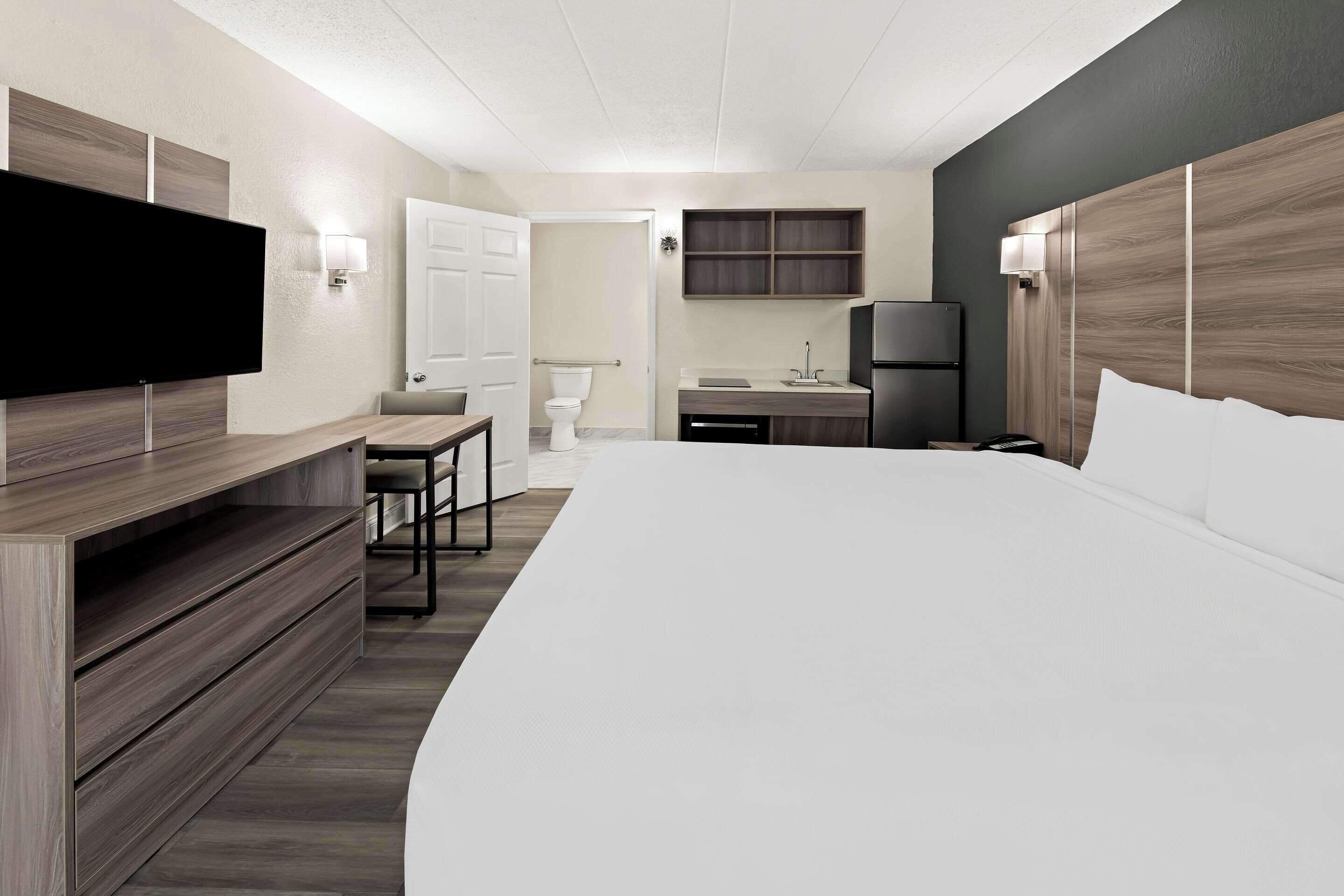 A modern hotel room with a large bed, a desk, a TV, and a small kitchenette.