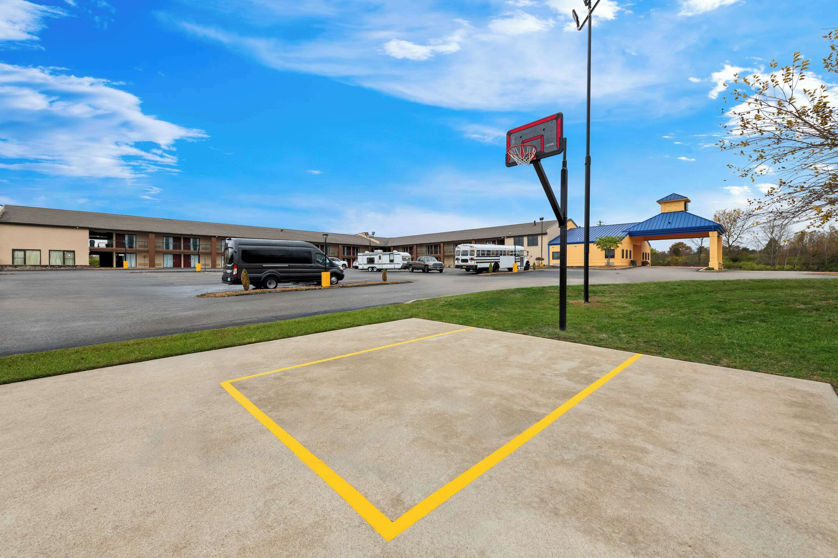 A basketball court with a hoop, a parking lot, and a building with a blue roof in the background.