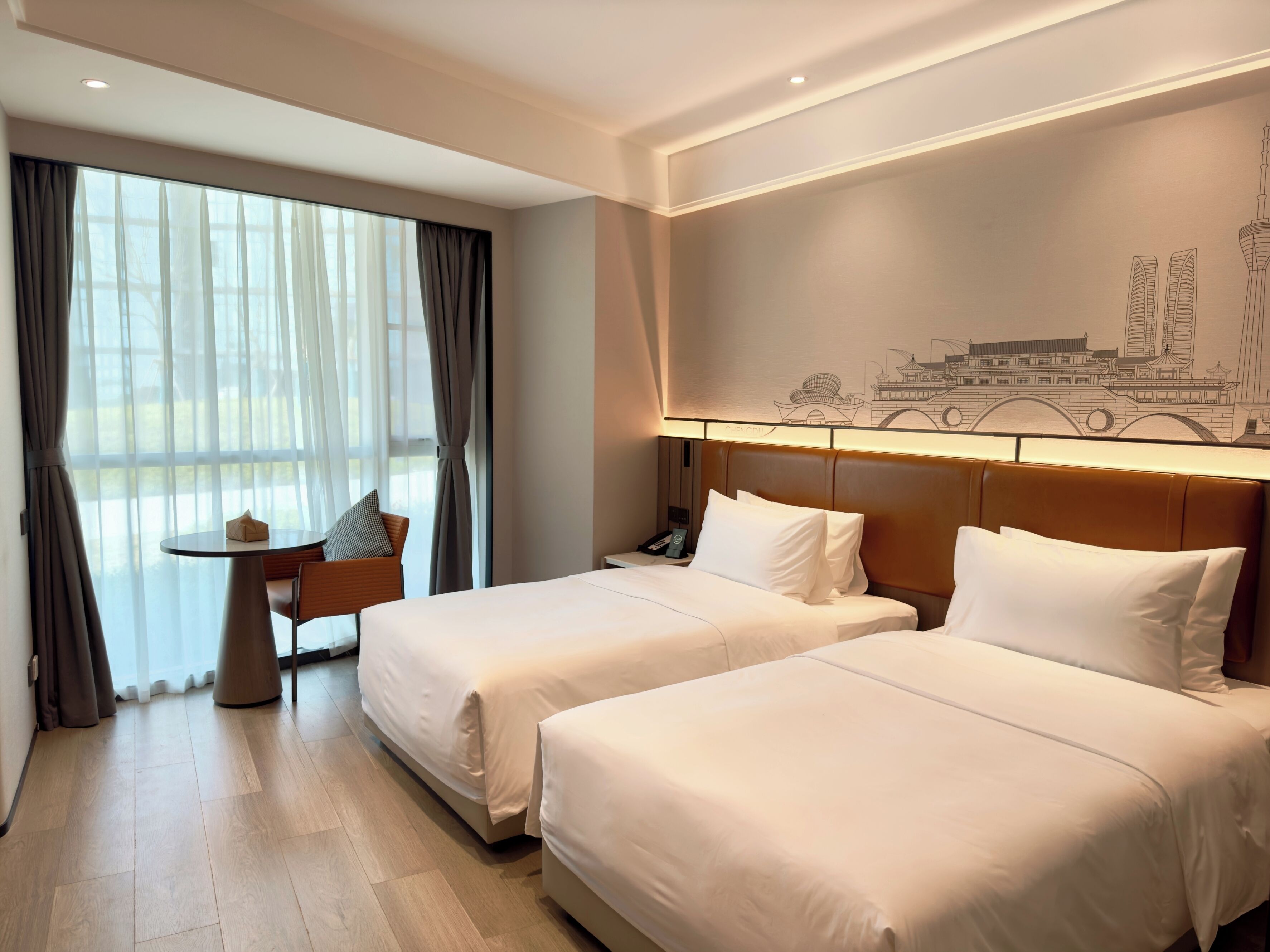 Ein Hotelzimmer mit zwei Betten, einem kleinen Tisch mit einem Stuhl und einem großen Fenster mit Vorhängen.