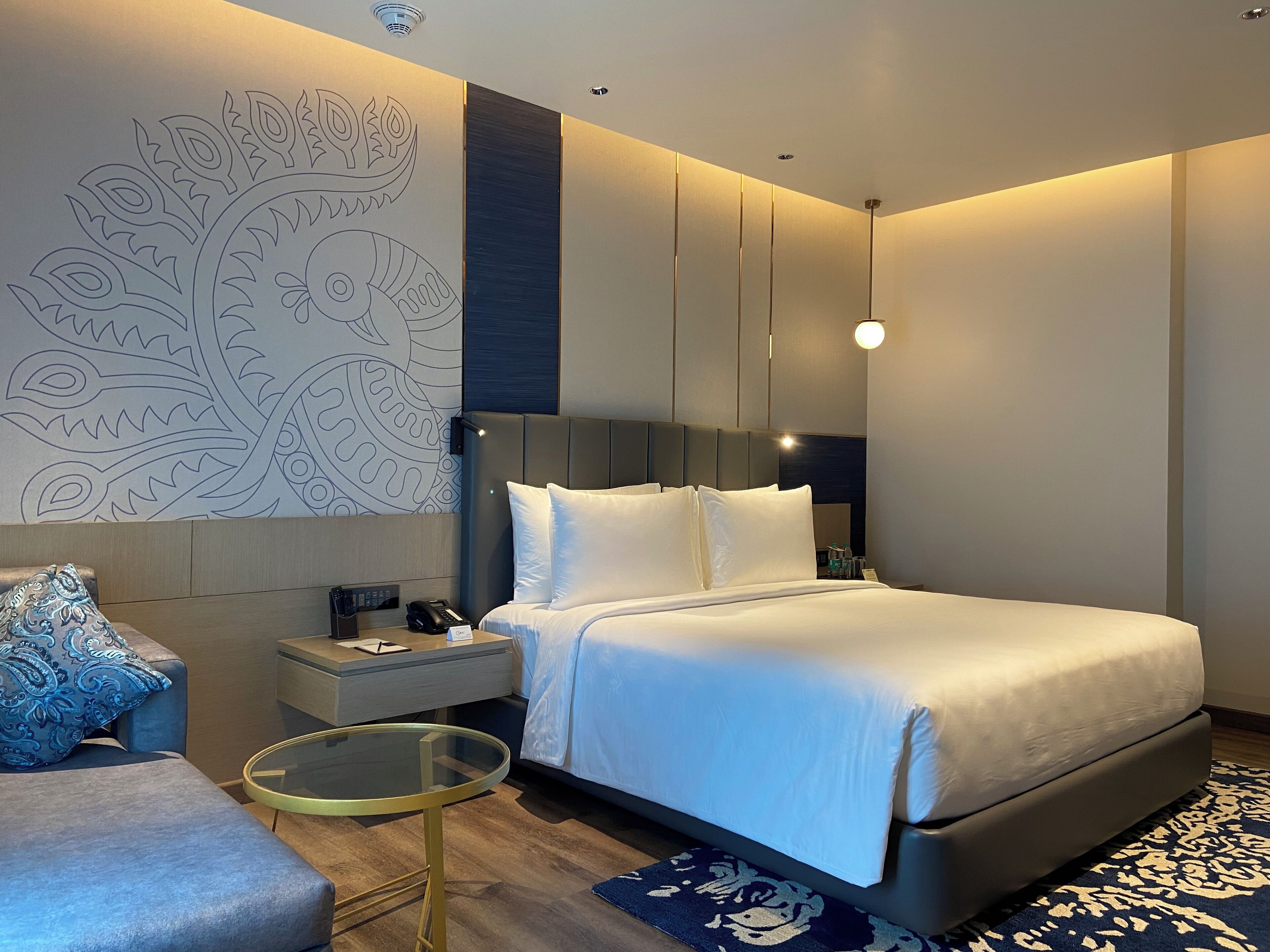 Una habitación de hotel moderna con una cama grande, una mesita de noche con un teléfono y una lámpara, una pequeña mesa redonda y un adorno de pared decorativo.