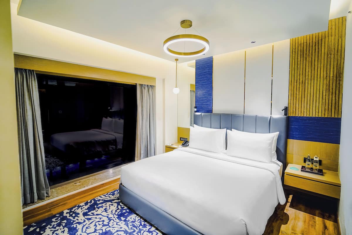 Een moderne hotelkamer met een groot bed, een nachtkastje met telefoon en fles, en een groot raam met gordijnen.