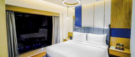 A modern hotel room with a large bed, a bedside table with a phone and a bottle, and a large window with curtains.
