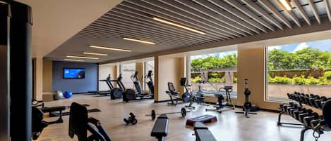 Un gimnasio moderno con diverso equipo de ejercicio, un televisor y grandes ventanales con vistas a zonas verdes.