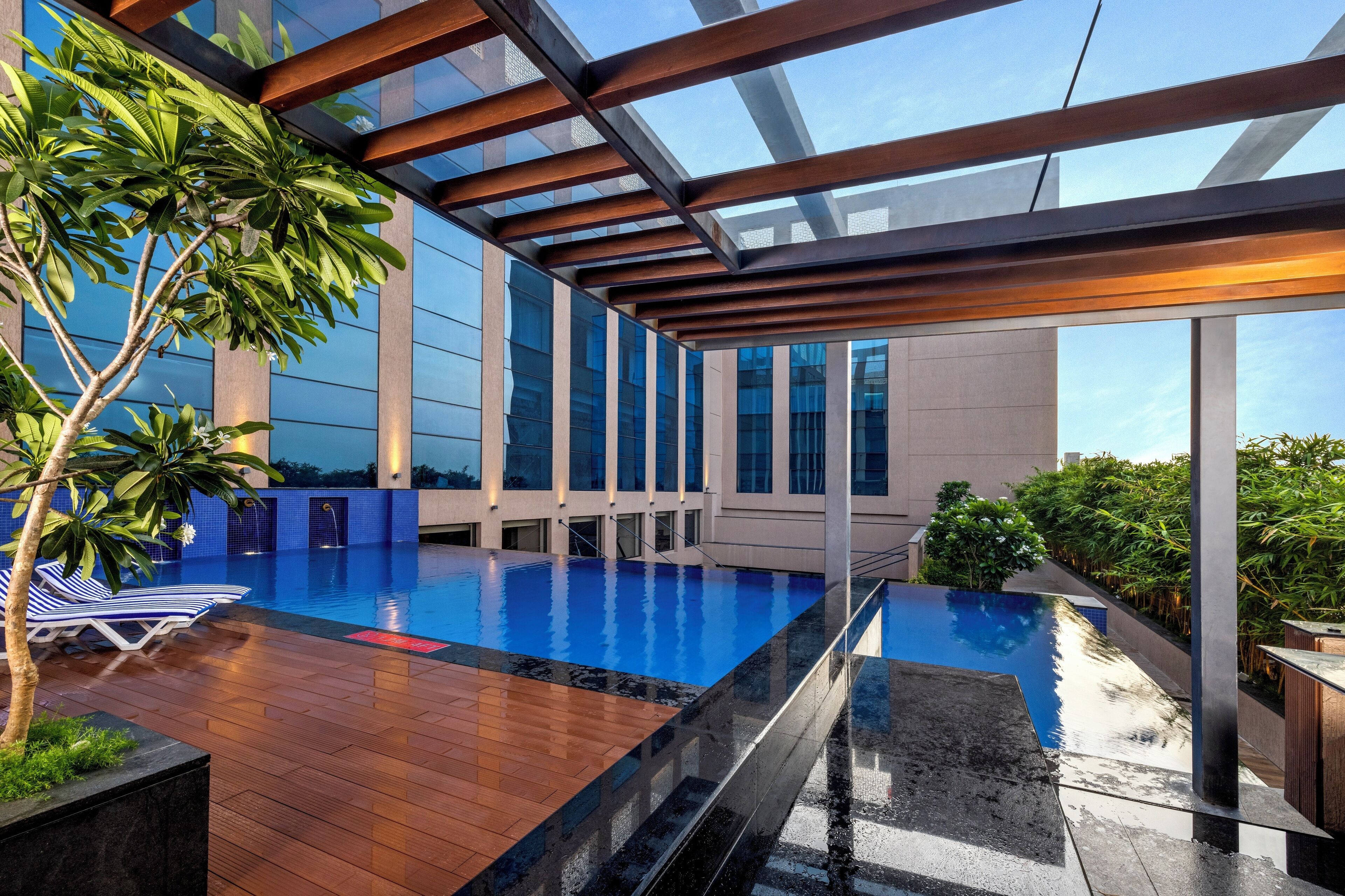 An infinity pool with wooden deck and glass railings, surrounded by lush greenery and modern architecture.