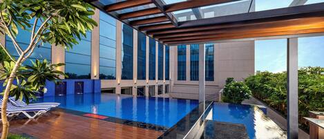 An infinity pool with wooden deck and glass railings, surrounded by lush greenery and modern architecture.