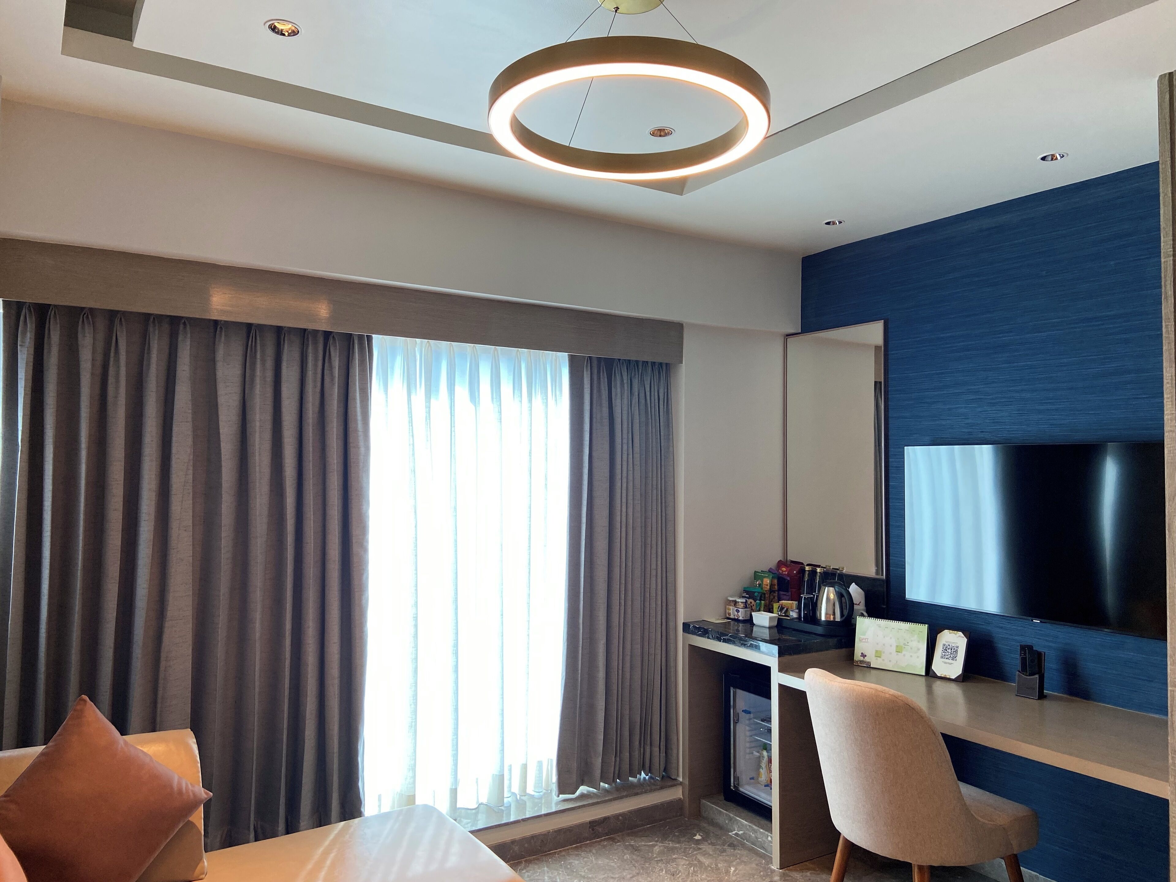 A modern hotel room with a large bed, a desk with a computer, a TV, and a minibar.