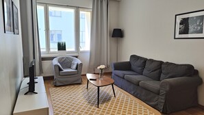 A modern living room with a grey sofa, a grey armchair, a small wooden table with a vase of white flowers, and a floor lamp. - Kotimaailma Aparthotel Kamppi 1BR with balcony (Helsinki)