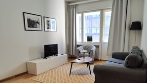 A modern living room with a flat-screen TV on a white console, a grey sofa, a small round table with a vase, and a patterned rug. - Kotimaailma Aparthotel Kamppi 1BR with balcony (Helsinki)