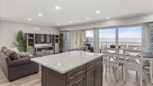 A modern kitchen with a marble countertop, a dining area with white chairs, and a view of the sea.