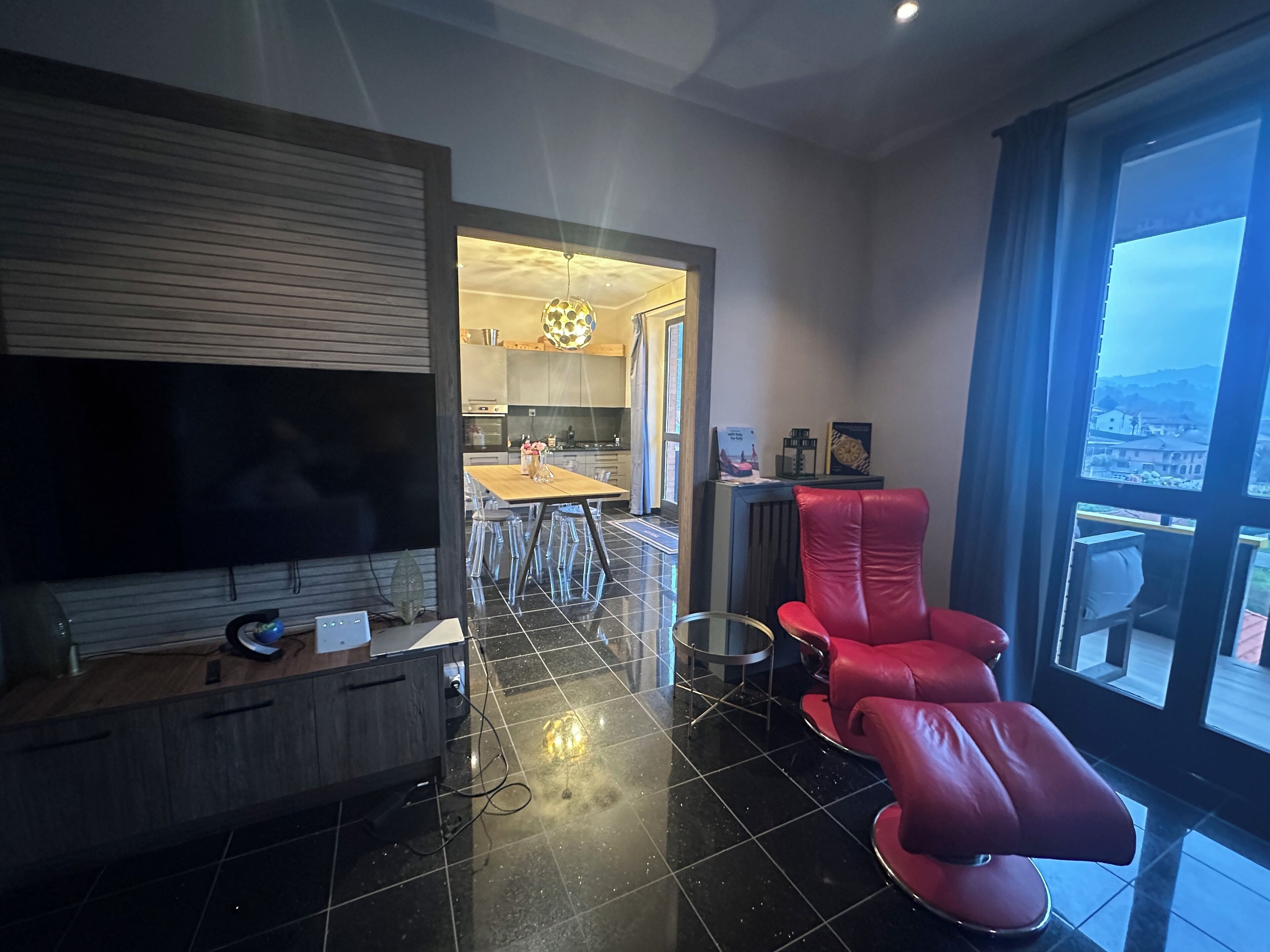 Een moderne woonkamer met een flatscreen televisie, een houten console, een rode leren fauteuil en een uitzicht op een keuken en eetruimte door een groot raam.