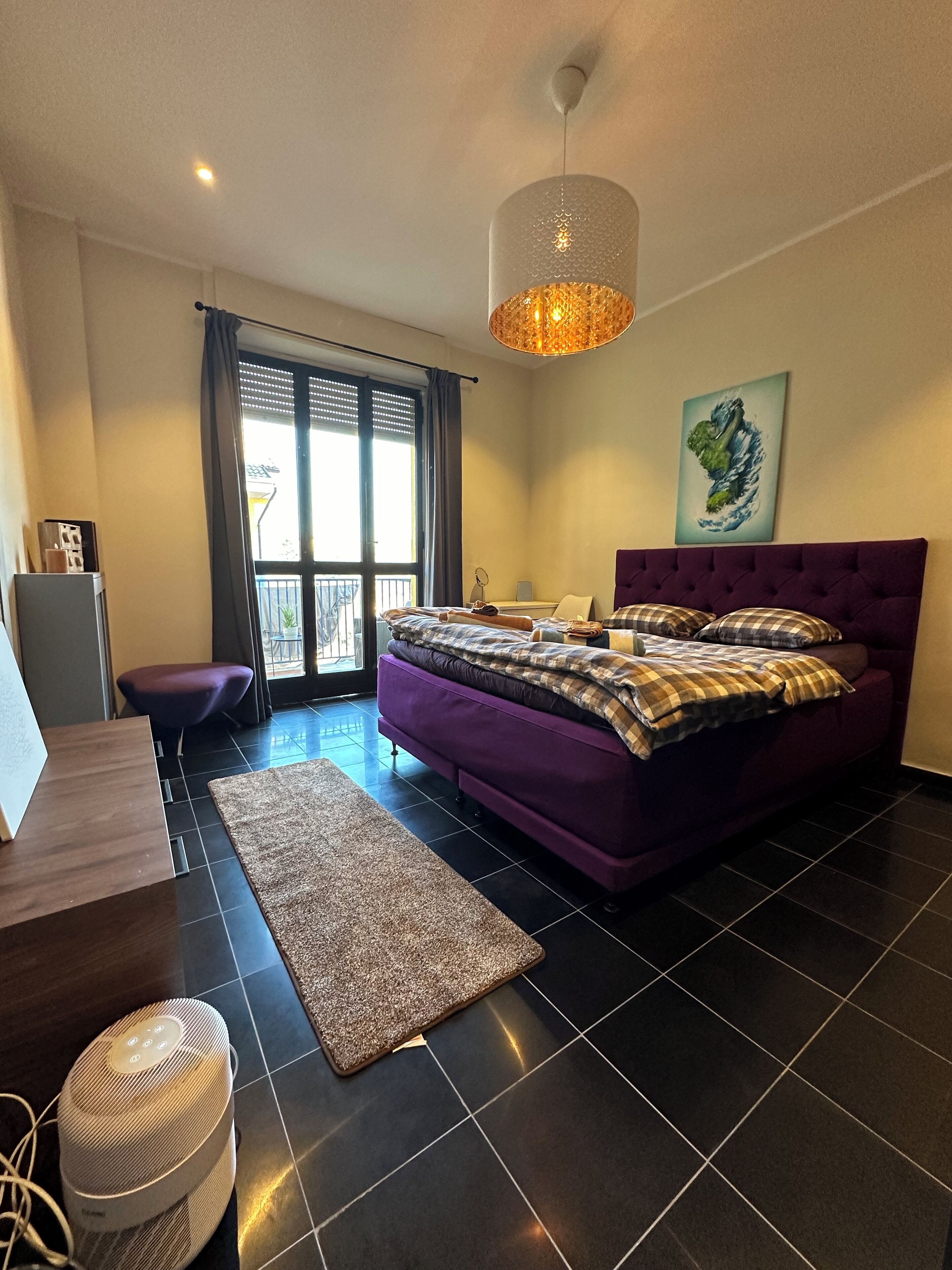 Un dormitorio moderno con una cama morada, un ventanal con persianas, una lámpara de pie y un cuadro en la pared.