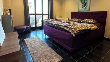 Un dormitorio moderno con una cama morada, un ventanal con persianas, una lámpara de pie y un cuadro en la pared.