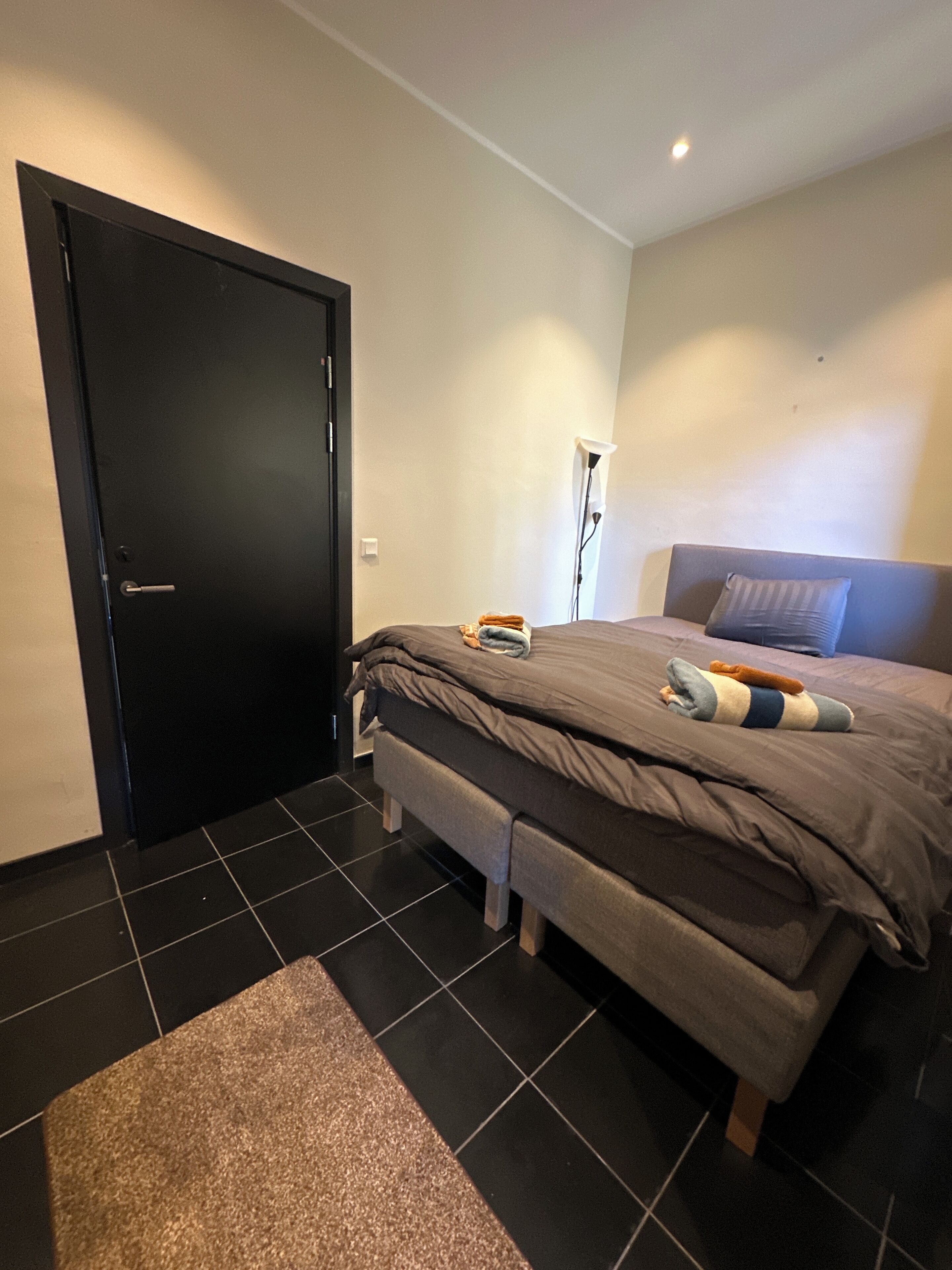 Une chambre à coucher moderne avec une porte sombre, un lit recouvert d’une couette grise, une lampe de chevet et un petit banc.