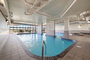 An indoor swimming pool with clear water, tiled floor, and a view of the sky through large windows.