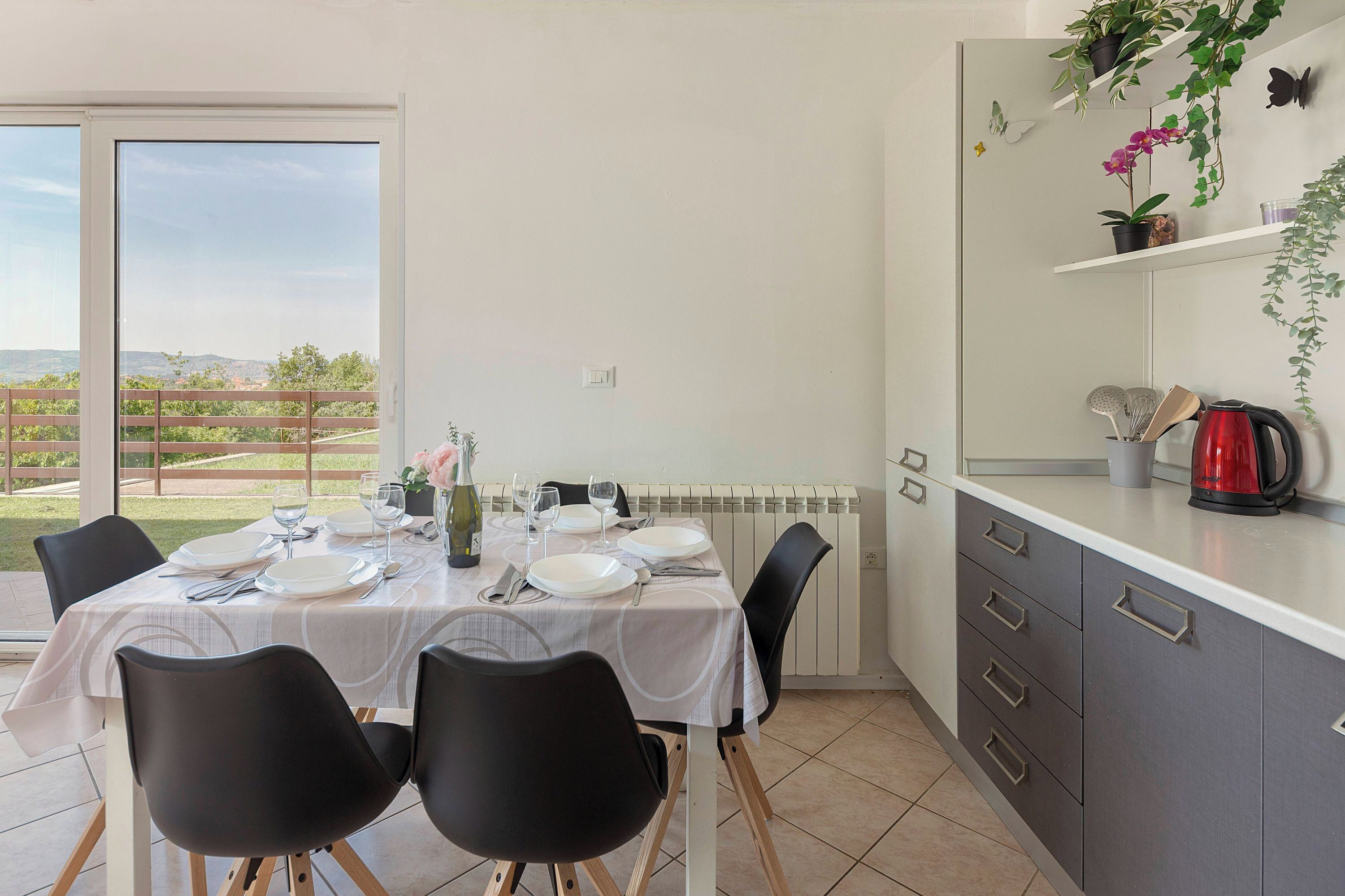 Un comedor con una mesa dispuesta para cuatro personas, una encimera de cocina con una tetera y vistas a un jardín a través de un ventanal.
