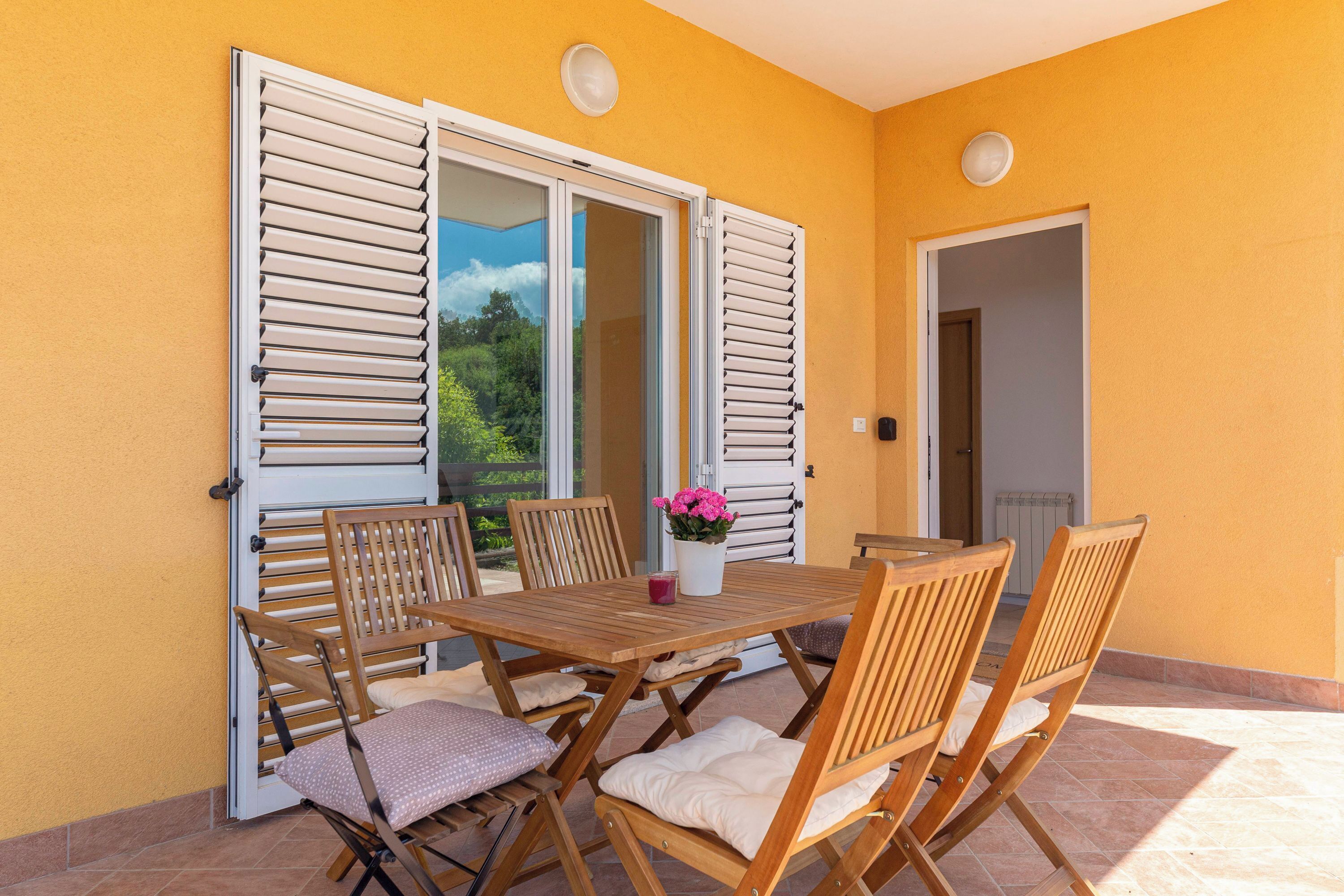 An outdoor patio with a wooden table and chairs, a white door, and a view of greenery.