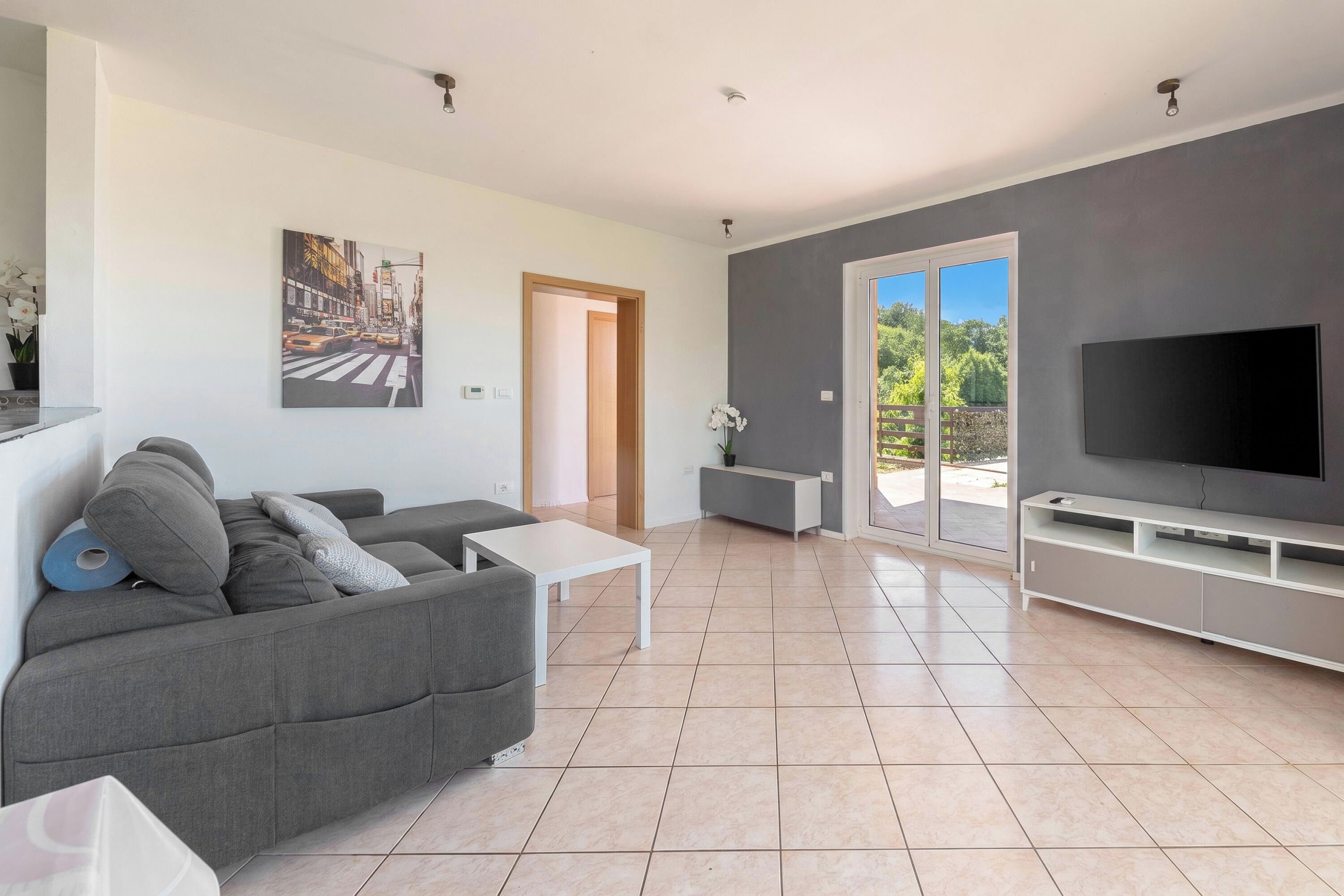 A modern living room with a grey sofa, a white coffee table, a flat-screen TV, and a sliding glass door leading to an outdoor area.