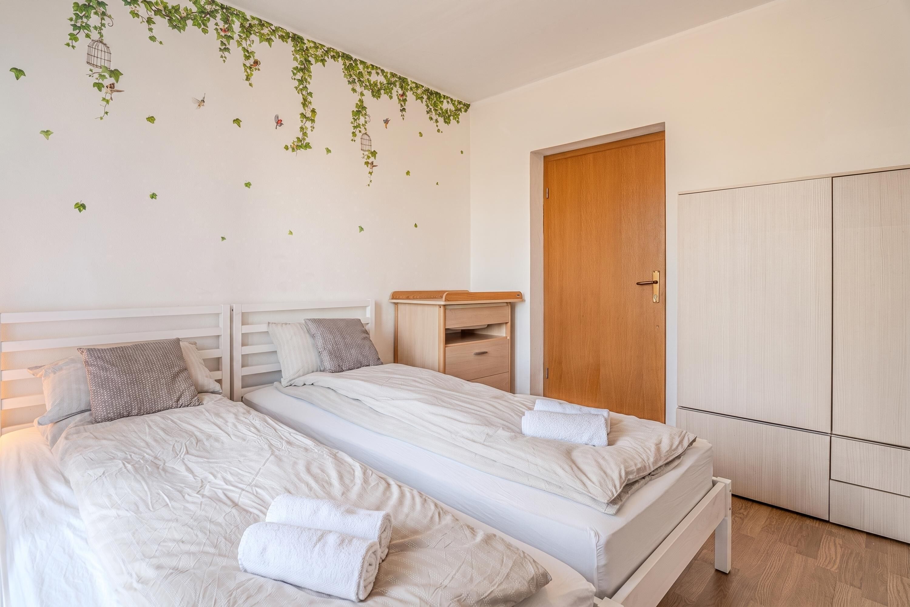 A bedroom with two beds, a wooden wardrobe, a desk, and a wall with green leaf decals.