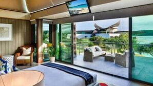 Quarto de hotel moderno com uma cama grande, área de estar com vista para a praia e uma tela de televisão.