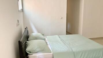 A single bed with a white mattress and a light green bedspread in a room with white walls and a door leading to another room.