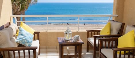 Un balcón con vista al mar, amueblado con dos sillas de madera y una mesita, además de un sofá con cojines.