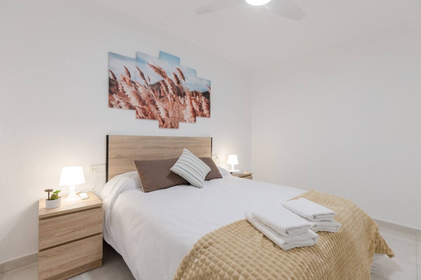 Un lit bien fait, avec une tête de lit, deux tables de chevet avec des lampes, une œuvre d’art accrochée au mur et un ventilateur de plafond.