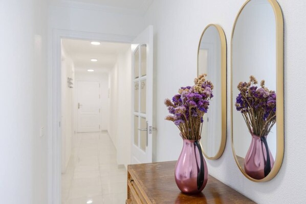Um corredor com uma mesa de madeira, dois vasos com flores secas e dois espelhos na parede.