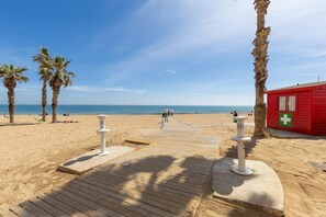 A beach with a wooden walkway leading to a red building with a green cross symbol, palm trees, and the ocean in the background. - Feel at Home by Fidalsa (La Mata)
