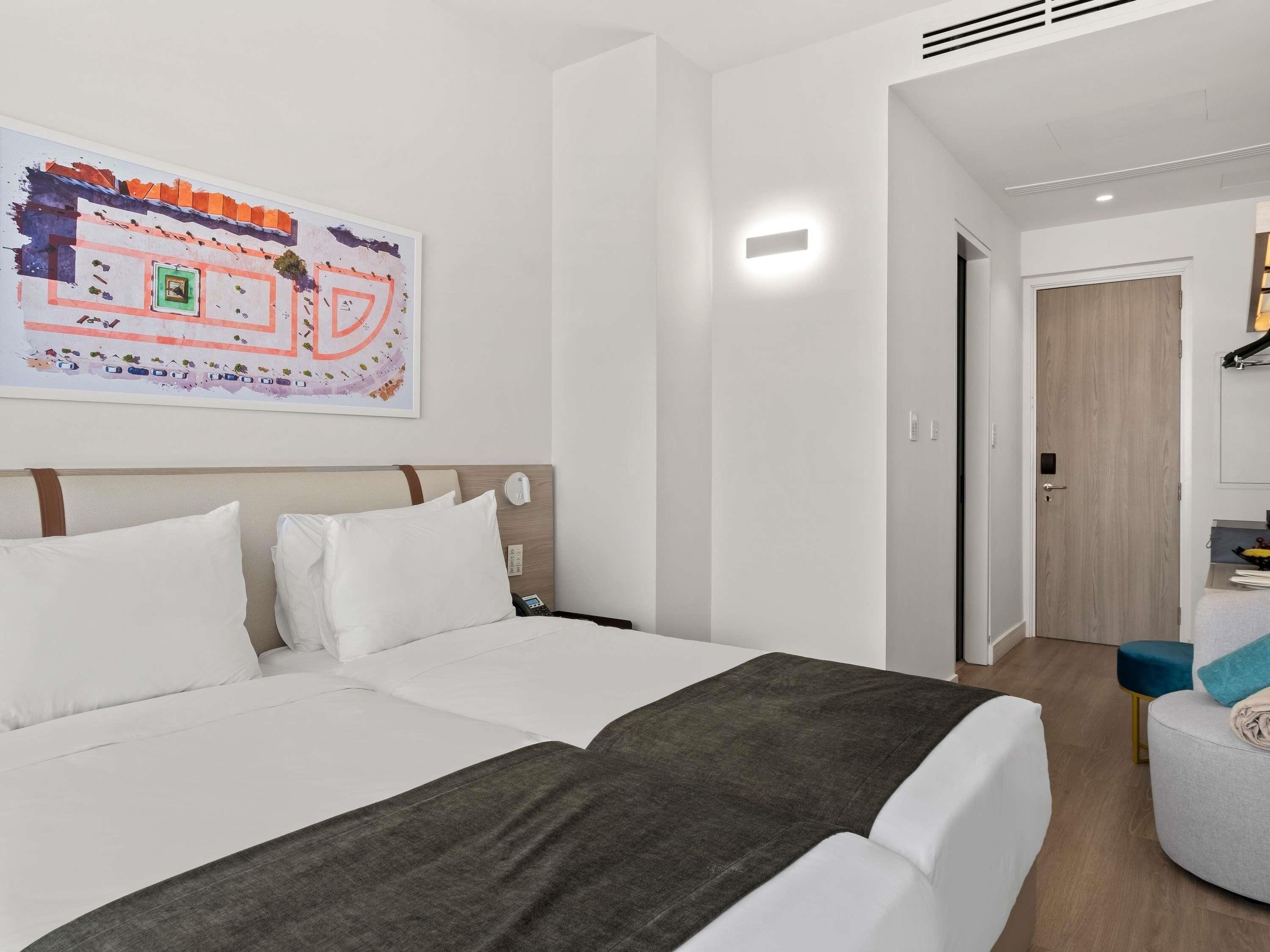 A hotel room with a bed, a bedside table, a nightstand, a wall-mounted lamp, a door, and a small seating area with a chair and ottoman.