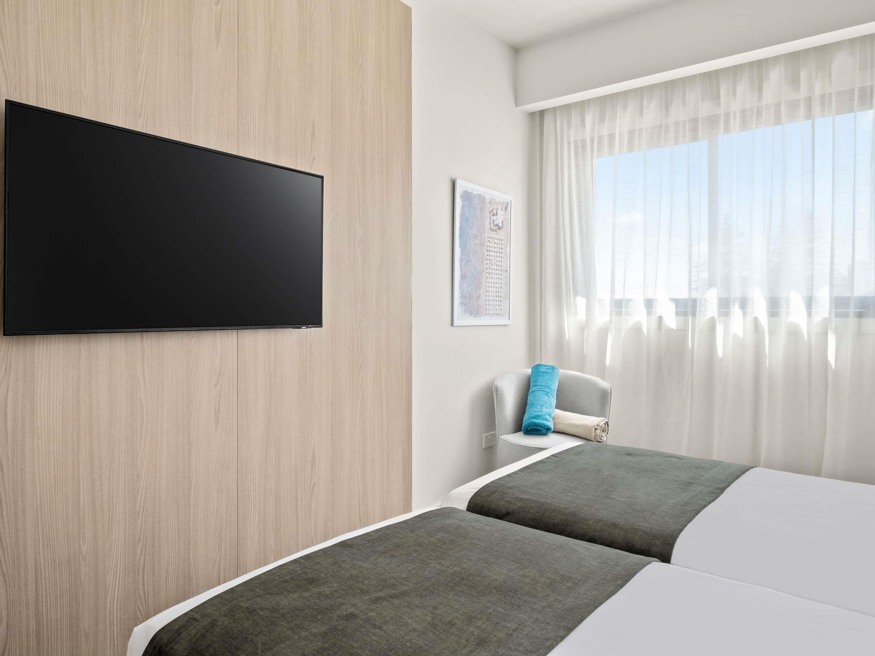 A hotel room with two beds, a flat-screen TV mounted on the wall, a window with sheer curtains, and a small chair with a blue towel.