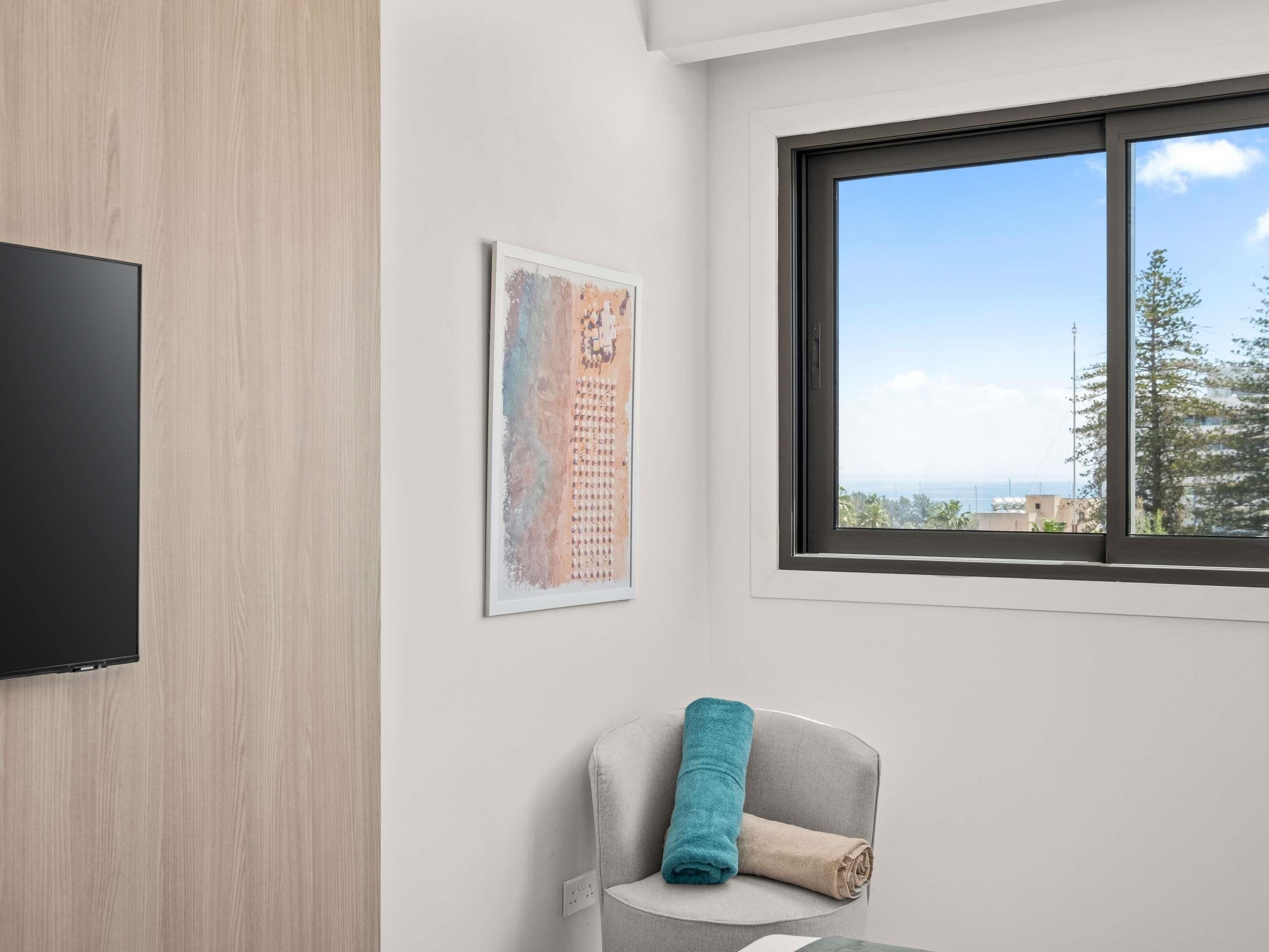 A modern room with a flat-screen TV, a painting, a window with a view, and a chair with a towel.