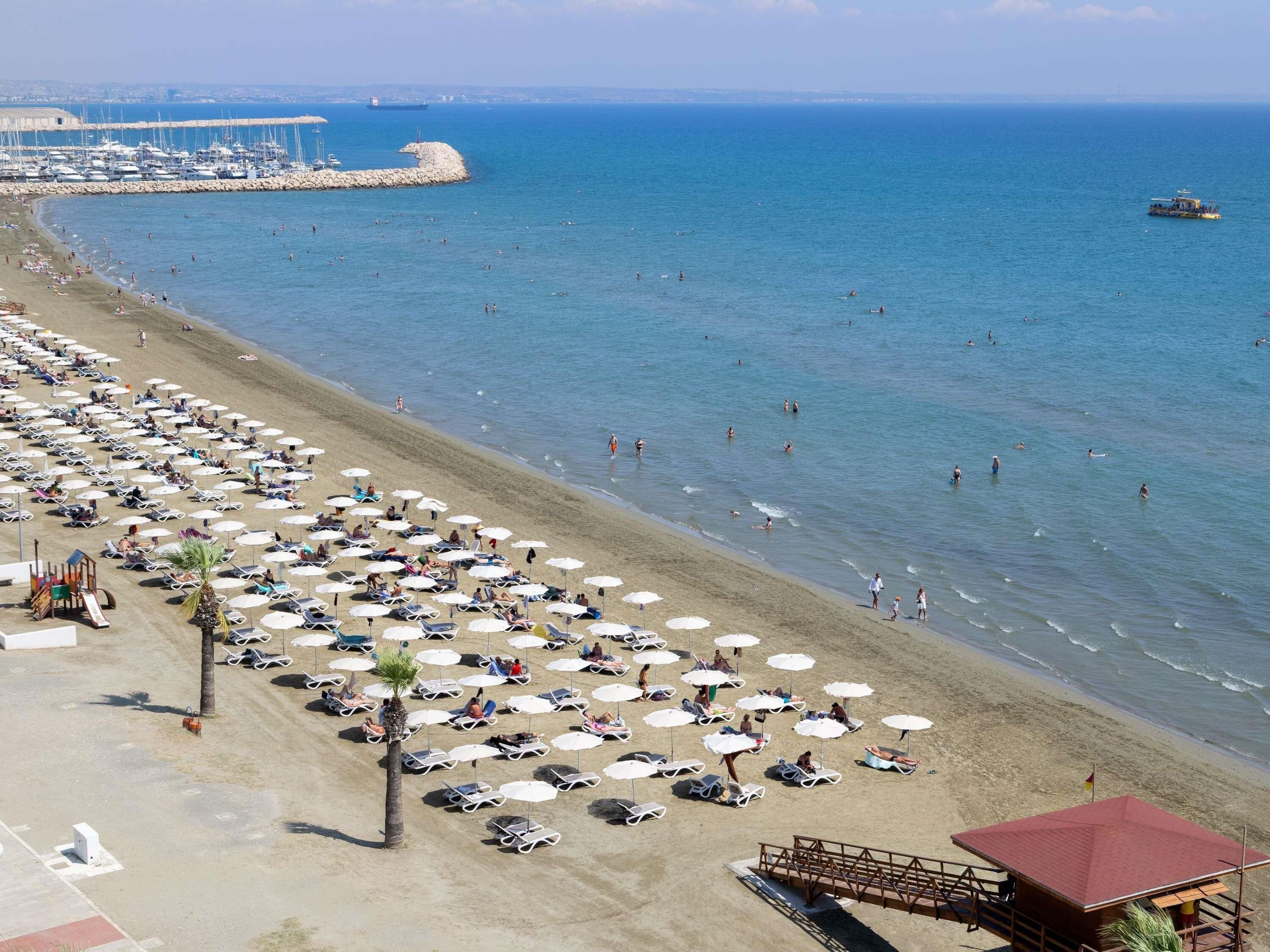 A beach with numerous sun loungers and umbrellas, a lifeguard tower, and a view of the sea and boats in the distance.