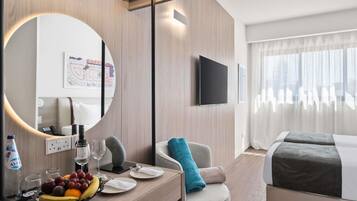 A modern hotel room with a wooden desk, a round mirror, a flat-screen TV, a bed with a grey and white duvet, and a large window with sheer curtains.