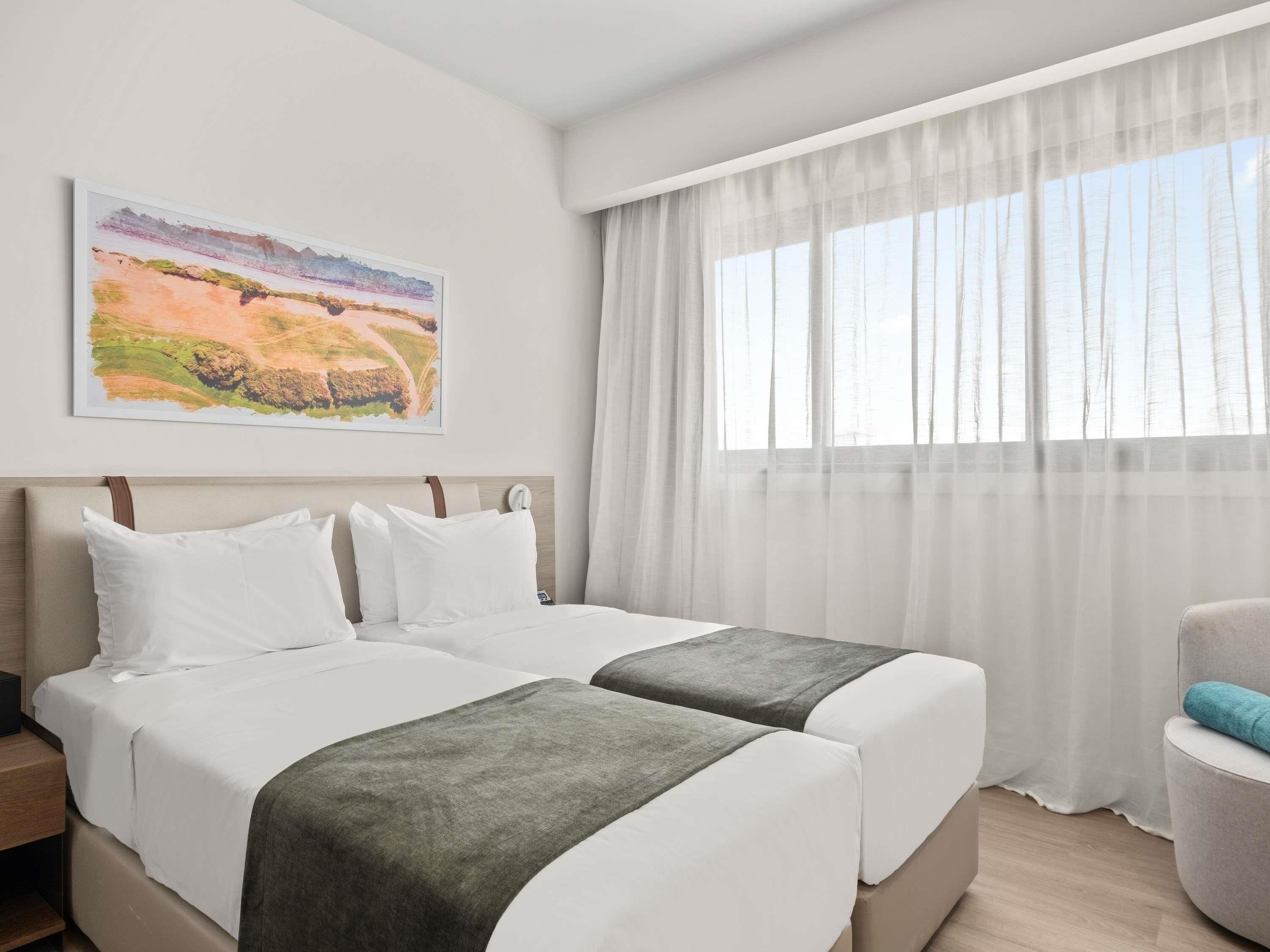 A hotel room with two beds, a large window with sheer curtains, a painting on the wall, and a chair.