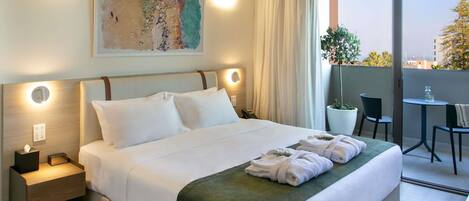 A hotel room with a bed, bedside tables, a painting, a balcony with a table and chairs, and a view of the city.