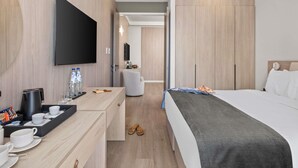 A modern hotel room with a bed, a flat-screen TV, a desk with a coffee maker, and a seating area with a chair and a small table. - Mercure Larnaca Finikoudes Beach (Larnaca)