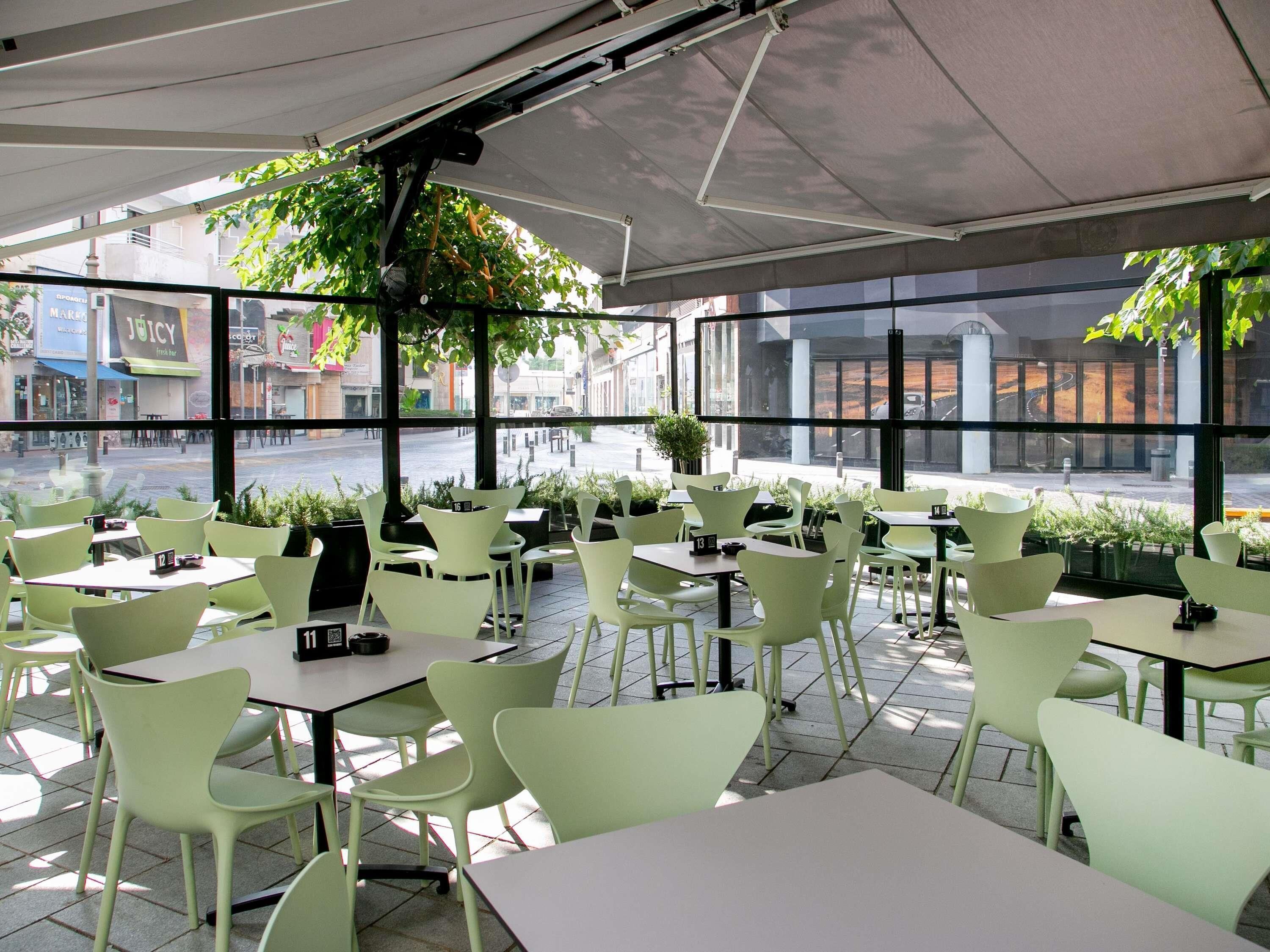 An outdoor cafe with green chairs and tables, large windows, and a view of the street.