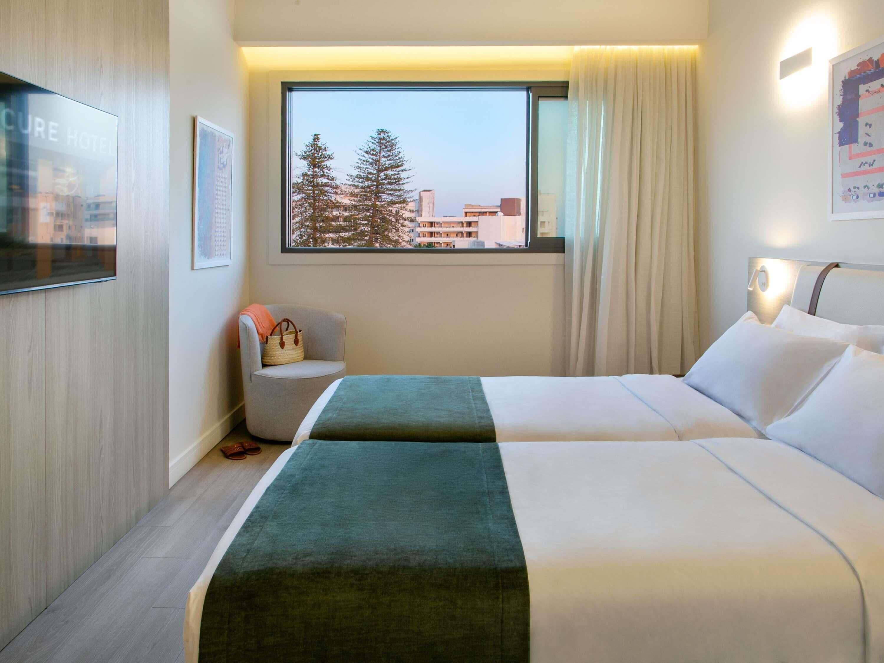 A hotel room with two beds, a chair, a window with a view of buildings and trees, and a mounted TV.