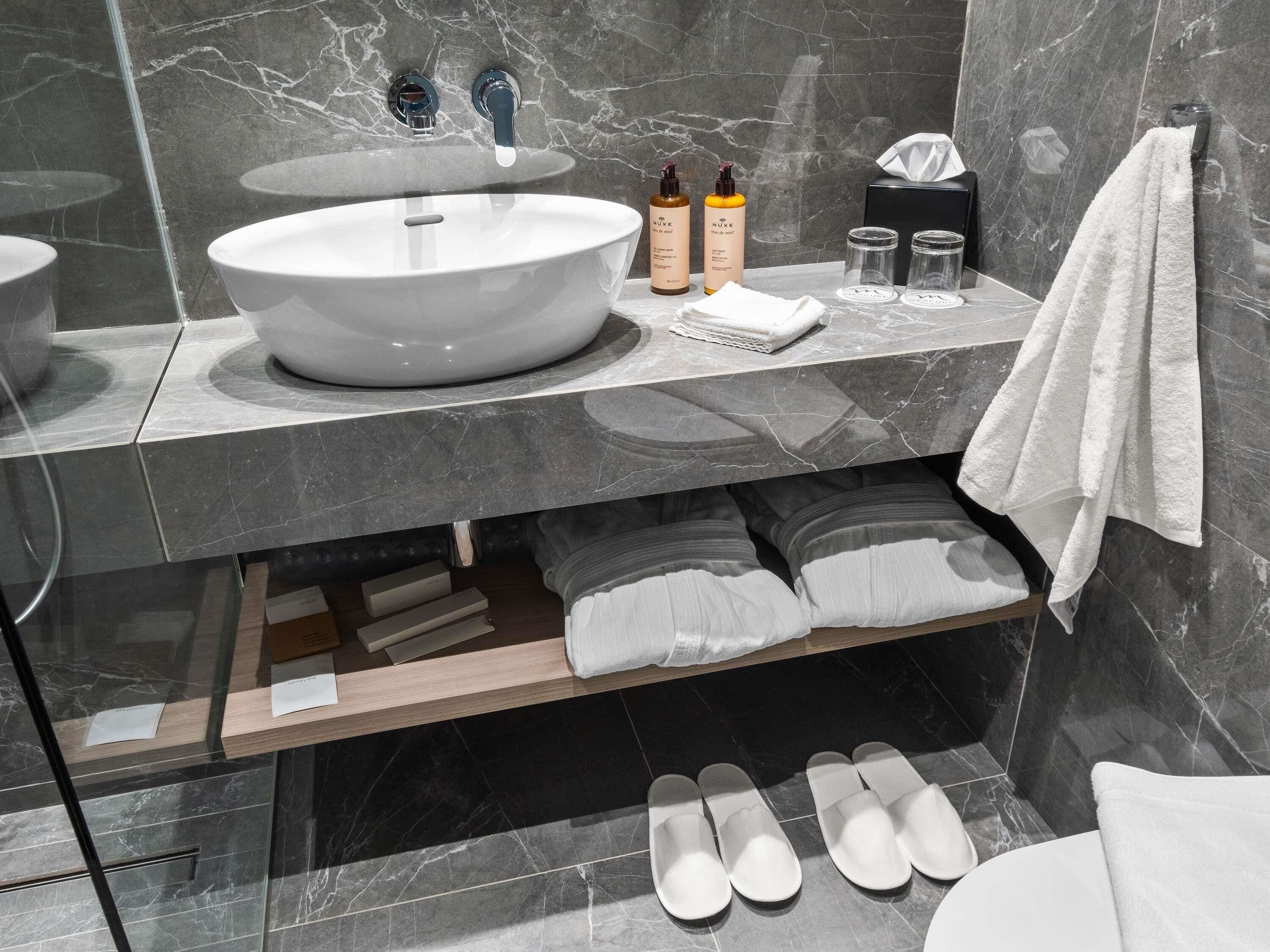 Une salle de bain moderne avec un grand lavabo ovale, deux robinets, un miroir, des articles de toilette, un porte-serviettes et une étagère avec des serviettes pliées et des chaussons.