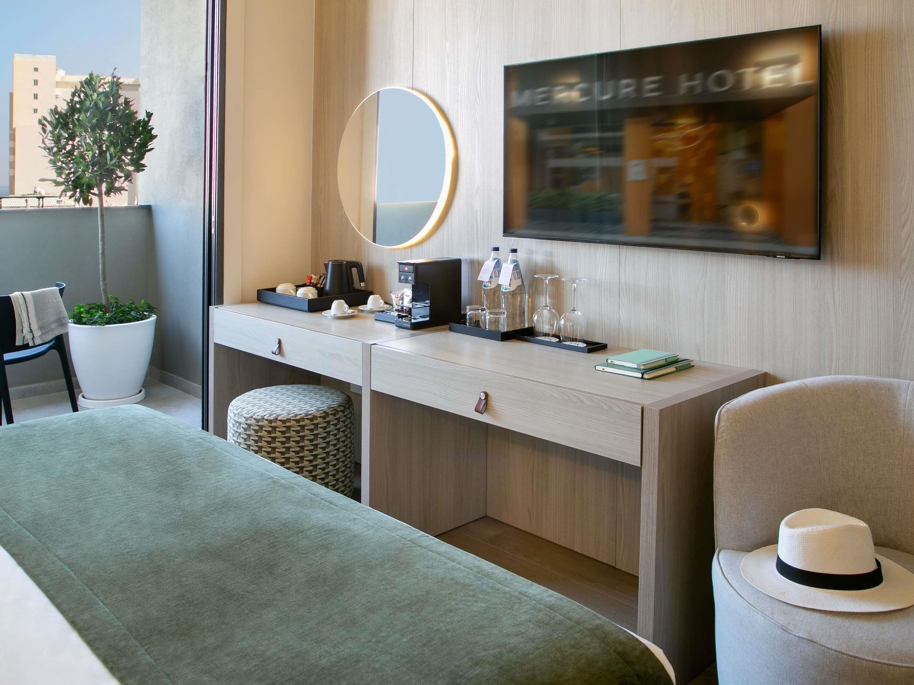 Habitación de hotel con cama, escritorio con espejo, cafetera y televisor que muestra 'MERCURE HOTEL'.